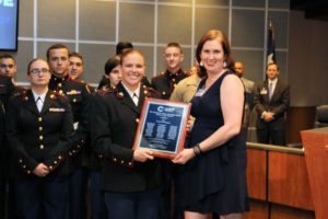 A woman in a black dress presents a plaque to a female Marine Corps cadet in a Board Room.