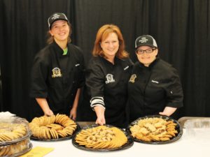 Three women hold trays of cookies.