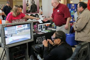 two men interact with a woman at a booth while a teenager drives a video game simulation.