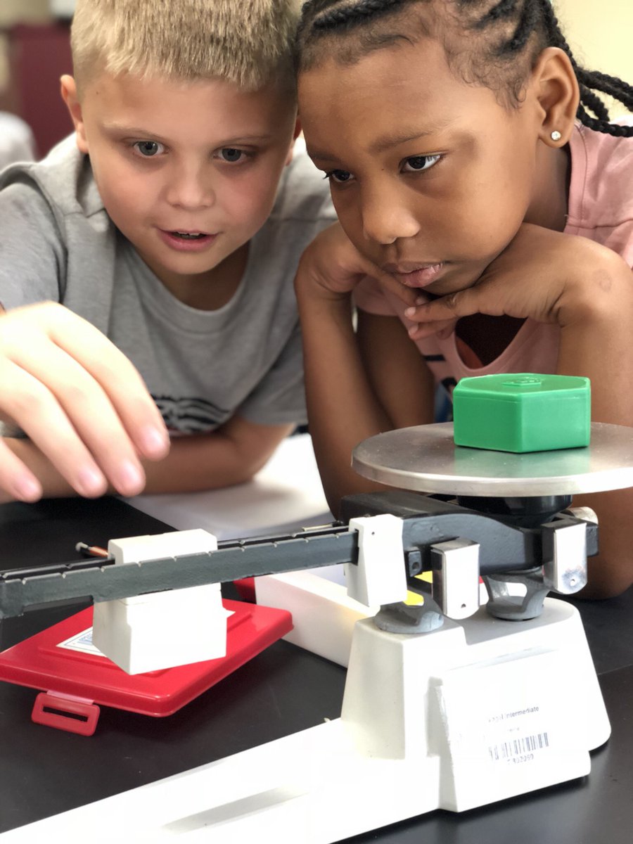 A boy and girl work together taking the mass of an item on triple beam balance.