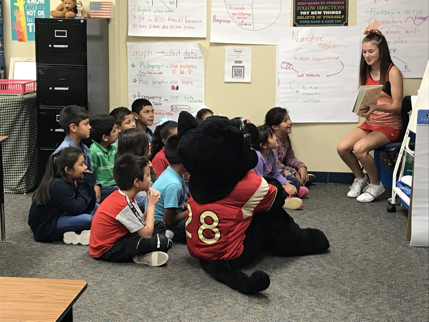 "a cheerleader and a school mascot read to a group of students"