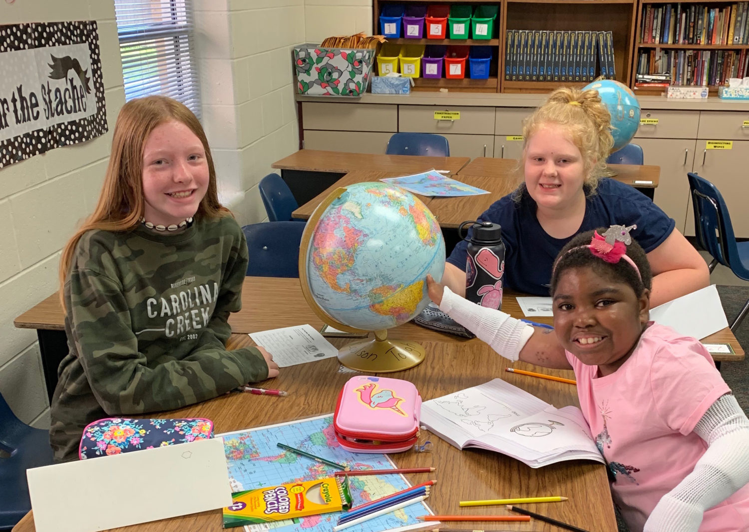 "three students sit at a desk with a world globe"