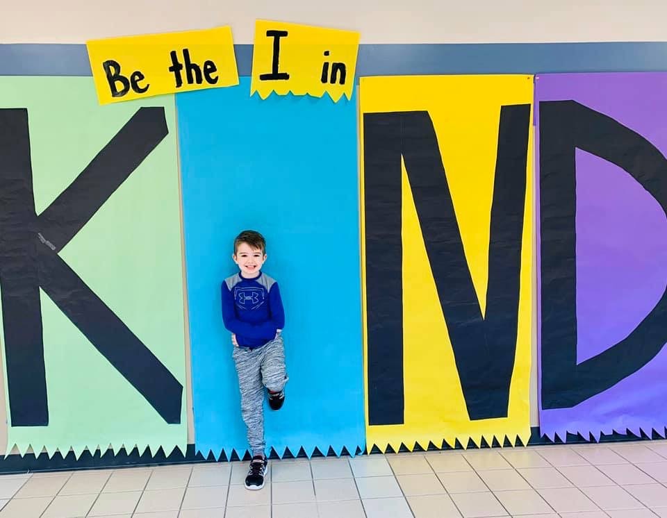 "a student poses in front of a mural the reads Be the I in Kind"