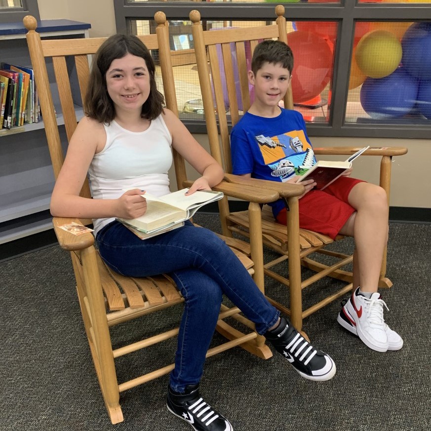 Two students smile for the camera while reading in rocking chairs.