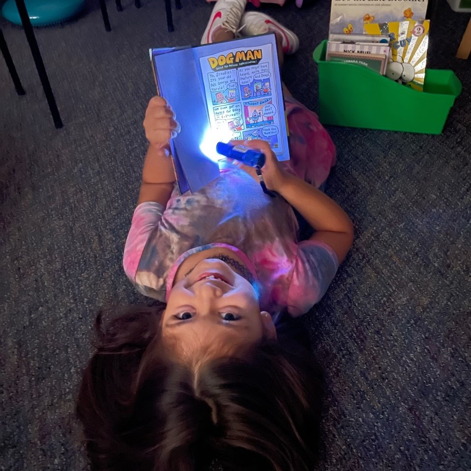 A student smiles while using a flashlight to read a book.