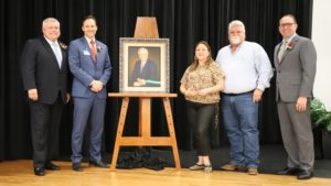 Four men and a woman stand on a stage around a portrait of a man.