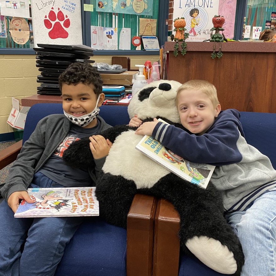 Two students smile while reading with a stuffed animal.